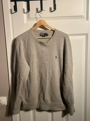 Grå sweatshirt från Polo Ralph Lauren - Säljer en stilren grå sweatshirt från Polo Ralph Lauren i bomull. Tröjan har rund halsringning, långa ärmar och den klassiska lilla loggan broderad på bröstet. Perfekt för en avslappnad och snygg look.