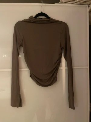 Brun långärmad topp - Säljer en brun långärmad polotopp med draperad effekt i midjan. Toppen har en smal passform och är tillverkad i mjukt, stretchigt material som sitter snyggt på kroppen. Perfekt att styla med jeans eller kjol för en cool look.