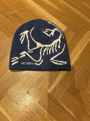 Blå mössa från Arc'teryx - Säljer en blå mössa från Arc'teryx med vit grafisk dinosaurie-design på framsidan. Mössan är ribbstickad och har en mjuk och skön känsla, perfekt för kalla dagar. Loggan är broderad i vitt framtill.