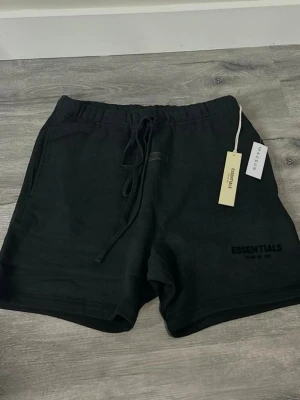 Svarta Essentials shorts med snörning - Säljer ett par svarta shorts från Essentials med snörning i midjan och tryckt logga på benet. De har en loose passform och är gjorda i mjukt bomullsmaterial, perfekta för chill dagar eller träning. Enkla att matcha med hoodies eller t-shirts.
