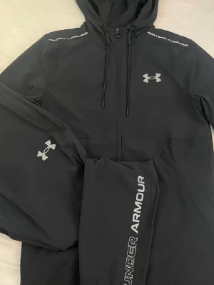Svart träningsset från Under Armour - Säljer ett svart träningsset från Under Armour med hoodie och matchande byxor. Setet har snygga loggor i silver på både jacka och byxa samt dragkedja och snörning på hoodien. Materialet är syntet och passar perfekt för träning eller chill.