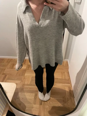Grå stickad v-ringad tröja med krage - Mysig grå stickad tröja med v-ringning och bred krage. Tröjan har långa ärmar, ribbade muddar och slits på sidorna för en chill och loose passform. Perfekt för lager-på-lager under höst och vinter. Säljes i storlek M.