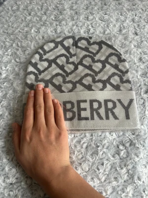  gråvit mössa med hjärtmönster - Snygg mössa i grått och vitt med coolt hjärtmönster och stor logga framtill. Mjuk och skön stickad kvalitet som håller dig varm. Perfekt accessoar för att lyfta din vinterstil.