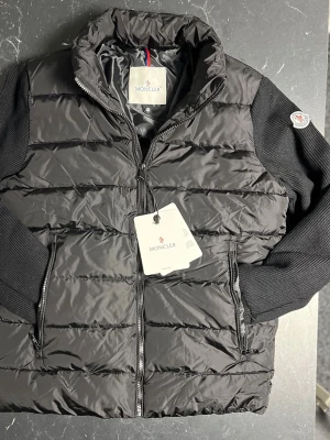 Svart monclair cardigan - Svart cardigan från Moncler med stickade svarta ärmar och klassisk logga på armen. Jackan har dragkedja framtill och två sidofickor med dragkedja. . Perfekt för dig som vill ha en snygg och varm jacka med street vibe.