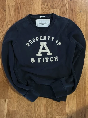 Mörkblå sweatshirt Abercrombie & Fitch - Säljer en lite schysstare mörkblå sweatshirt från Abercrombie & Fitch med stor vit broderad text och ett stort A på bröstet. Tröjan har rund hals, ribbade muddar och är långärmad. Perfekt för dig som gillar klassisk college-stil. Storlek L - PASSAR S/M