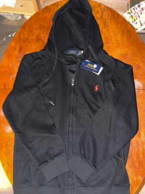 Ralph Lauren Zip Hoodie - Snygg svart zip hoodie  med dragkedja och huva. Klassisk röd logga broderad på bröstet, snörning i huvan och två praktiska fickor framtill. Perfekt för chill dagar och enkel att matcha med jeans eller joggers.
