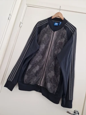 Adidas Originals herr träningsjacka 2XL - Snygg Adidas Originals träningsjacka i mörkgrå/svart med unikt mönstrat framparti. Klassiska tre ränder längs ärmarna och dragkedja hela vägen framtill. Storlek 2XL  I riktigt fint skick – inga fläckar eller skador. Enda lilla anmärkningen är att det grå partiet runt kragen har börjat flagna/ljusna lite, men det syns inte vid användning.   Detta är ett plagg som är riktigt svårt att få tag på idag – perfekt för samlare och adidas-fans!