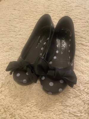 Svarta ballerinaskor med prickar och rosett - Säljer ett par svarta ballerinaskor från KX & Co. med vita prickar och stora svarta rosetter framtill. Skorna har rund tå och platt sula, tillverkade i syntetmaterial. Perfekt för dig som gillar lekfulla detaljer och mönster.