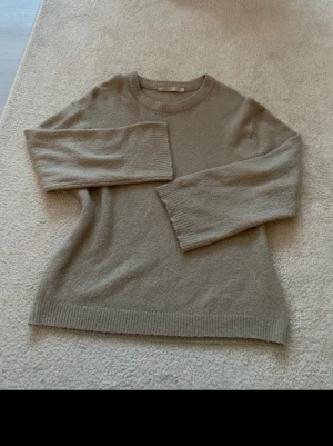 Beige stickad tröja med rund hals - Mysig beige stickad tröja med rund hals och långa ärmar. Tröjan har ribbade muddar vid ärmslut och nederkant, vilket ger en enkel men stilren look. Perfekt för att hålla värmen under kyliga dagar.
