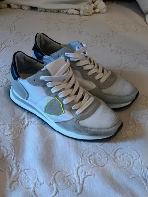 Vita och grå sneakers från Philippe Model - Snygga sneakers från Philippe Model i vitt och grått med detaljer i mocka och skinn. Skorna har en svart häl, vit sula och coolt neongult kantad logga på sidan. Klassisk snörning och platt sula. Perfekta för dig som gillar stilrena men ändå unika sneakers. passar bra nu till vintern 