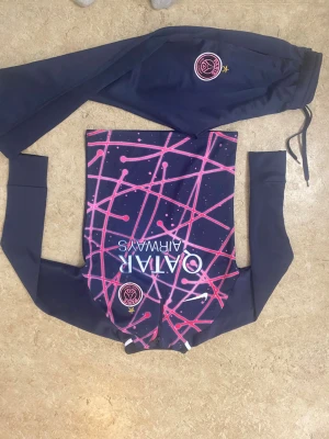 PSG träningsset i marinblått och rosa - Säljer ett fett PSG träningsset från Nike med långärmad tröja och byxor. Tröjan har ett galet mönster i rosa och lila på framsidan, PSG-logga och Qatar Airways-tryck. Byxorna är marinblå med PSG-märke och dragsko i midjan. Perfekt för dig som älskar fotboll och vill sticka ut på planen.