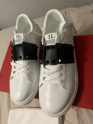Valentino Rockstud Untitled sneakers - Snygga vita Valentino Rockstud Untitled sneakers med svarta detaljer och ikoniska nitar över snörningen. Skorna har rund tå, platt sula och är tillverkade i skinn. Perfekt för dig som vill sticka ut med en lyxig och edgy stil.