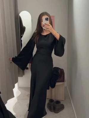 Svart satin maxiklänning med vida ärmar - Snygg svart maxiklänning i satin med vida ärmar och markerad midja. Klänningen har en elegant siluett och knytning i ryggen som ger en extra detalj. (HELT NY MED PRISLAPP)