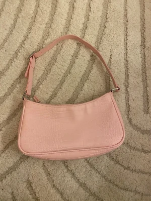 Rosa axelväska från Monki - Säljer en ljusrosa axelväska från Monki med croco-mönstrad yta i syntetmaterial. Väskan har justerbar axelrem och stängs med dragkedja. Perfekt storlek och super fin färg! iprincip oanvänd och super fint skick!!