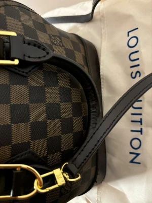 Alma bb - Inte äkta. Säljer en elegant Alma BB handväska i klassiskt Damier-mönster. Väskan är brun och svart med guldfärgade metalldetaljer, har två handtag och avtagbar axelrem. Insidan är klarröd.