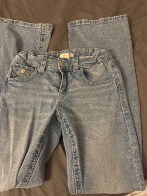 Ljusblå jeans med flare ben Gina  - Säljer ett par ljusblå jeans med klassisk femficksdesign och flare ben. Jeansen har knapp och dragkedja framtill samt bälteshällor. Materialet är jeans och färgen är snyggt tvättad för en avslappnad look. Storlek 164 från Gina young