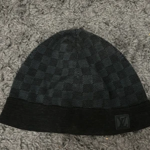 Svart rutig mössa från Louis Vuitton - Snygg svart mössa från Louis Vuitton med diskret rutigt mönster och LV-logga framtill. Mössan är stickad i mjuk ull och kashmir, vilket gör den både varm och skön. Perfekt accessoar för att lyfta din look under kyliga dagar.