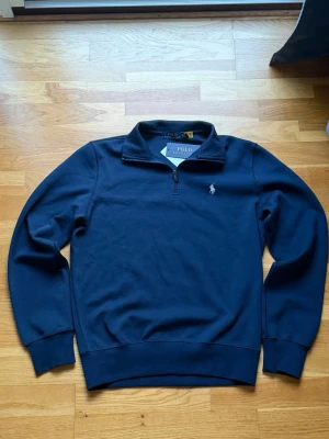 Marinblå half zip från Polo Ralph Lauren - Snygg marinblå tröja från Polo Ralph Lauren med half zip och klassisk broderad logga på bröstet. Tröjan har långärmad design, ribbade muddar och är gjord i mjuk bomull. Perfekt för dig som gillar stilrena och sportiga plagg.