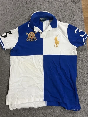 Blåvit rutig polotopp från Polo Ralph Lauren - Säljer en blåvit polotopp från Polo Ralph Lauren med rutigt mönster och broderade detaljer på bröstet. Toppen har korta ärmar, klassisk krage och stora siffror på ärmarna. Materialet är mjuk bomull och toppen har en slim passform. M/S