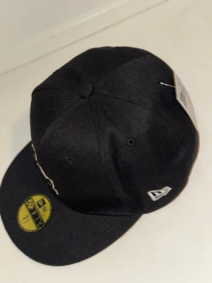 Svart LA 59FIFTY keps från New Era - Säljer en svart New Era 59FIFTY keps med LA-logga framtill i vitt och svart broderi. Kepsen har en klassisk rak skärm i grått och detaljer från Cooperstown Collection. Perfekt för dig som gillar streetwear och sportig stil.