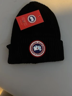 Svart ribbad mössa från Canada Goose - Svart ribbstickad mössa från Canada Goose med klassisk rund logotyp framtill. Mössan har uppvikt kant och är tillverkad i ett mjukt material som håller dig varm under kalla dagar. Perfekt för dig som vill ha en stilren och ikonisk accessoar.