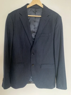 Mörkblå randig kavaj från Riley - Snygg mörkblå kavaj från Riley med smal passform och tunna vita ränder. Kavajen har två knappar framtill, klassisk krage och tre dekorativa knappar vid ärmslutet. Materialet är slätt och känns lyxigt, perfekt för dig som gillar stilrena detaljer.