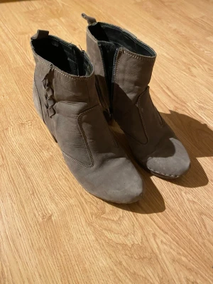 Gråa/bruna boots från Emma, storlek 39 - Säljer ett par snygga gråa boots från Emma i storlek 39. Skorna har en låg klack, rund tå och är gjorda i mjukt skinn. På sidan finns en flätad detalj och dragkedja för enkel på- och avtagning. Perfekt för dig som gillar stilrena och bekväma skor.