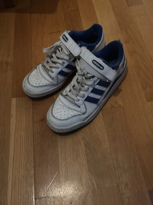 Adidas sneakers vita och blå - Säljer ett par klassiska Adidas sneakers i vitt och blått med de ikoniska tre ränderna på sidan. Skorna har snörning och kardborreband upptill för extra support. Ovandelen är i skinn och insidan är fodrad i blått tyg. Perfekta för dig som gillar retrostil.