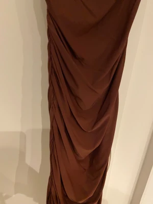 Brun långklänning - Säljer en snygg brun långklänning från Shein med tunna axelband och draperad framsida. Klänningen har figurnära passform och är tillverkad i ett mjukt, stretchigt material som ger en elegant siluett. Perfekt för dig som vill sticka ut med en stilren look. Man får dessutom en väldigt fin passform, rekommender😻(använd 2 gånger tidigare)