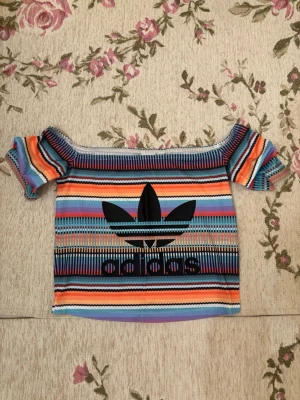 Randig offshoulder topp från Adidas - Säljer en färgglad offshoulder topp från Adidas med ikonisk svart logga framtill. Toppen har ett randigt mönster i orange, blått, lila, svart och vitt. Materialet är mjukt och stretchigt, perfekt för dig som gillar sportig och unik stil.