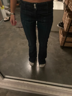 Mörkblå bootcut jeans från Ginatricot - Säljer ett par mörkblå jeans från Ginatricot med bootcut passform och hög midja. Jeansen har två stora fickor fram, dubbla knappar och dragkedja. Materialet är klassisk jeans med tydliga sömmar längs benen. Perfekt för dig som gillar retrovibbar och snygg siluett.