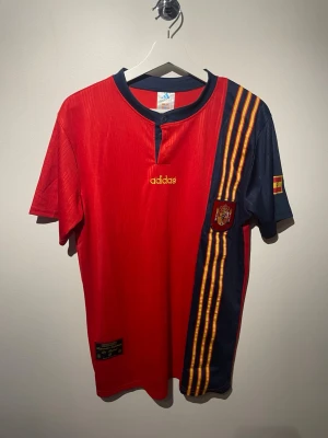 Retro spanien fotbollströja - Säljer en officiell röd Spanien fotbollströja från Adidas i storlek M. Tröjan har marinblå detaljer, gula ränder på ena ärmen och Spaniens landslagsemblem. Materialet är polyester och den har en klassisk rund hals med knapp. Perfekt för dig som älskar fotboll och landslagströjor.