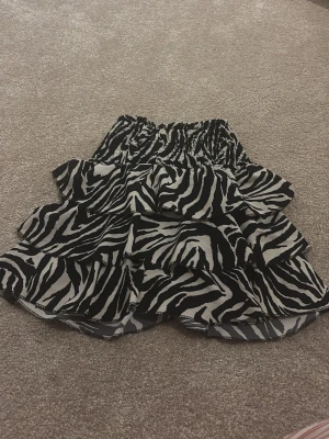 Zebramönstrad volangkjol - Säljer en kort kjol med zebramönster i svart och vitt. Kjolen har flera lager med volanger och en elastisk midja som gör den bekväm och enkel att bära. Perfekt för dig som gillar djurmönster och vill sticka ut lite extra.