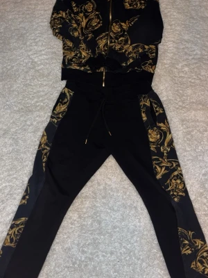 Versace Jeans Couture mjukisbyxor svart/guld - Säljer ett par svarta mjukisbyxor från Versace Jeans Couture med ikoniskt guldigt barockmönster längs sidorna. Byxorna har snörning i midjan och är tillverkade i mjukt bomullsmaterial. Perfekta för dig som vill ha en lyxig och bekväm look.