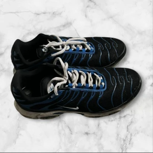 Nike Air Max Plus svarta och blå sneakers - Nike Air Max Plus sneakers i svart och blått med vita snören och TN-logga på hälen. Skorna har ett vågigt mönster i blått över ovandelen och en synlig Air Max-sula. Materialet är syntet och mesh, med rund tå och platt sula. Perfekt för dig som gillar sportig stil.