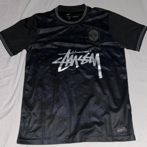 Svart Nike x Stüssy Brasil fotbollströja - Nike x Stüssy Brasil fotbollströja i svart med silverfärgat Stüssy-tryck på bröstet och broderat Brasil-märke. Tröjan har rund hals med ribbade detaljer och är tillverkad i lätt, ventilerande Dri-Fit polyester. Perfekt för dig som gillar streetwear och fotboll.