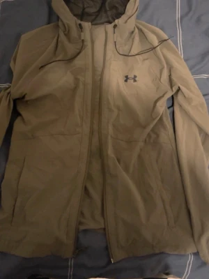 Under Armour vindjacka  - Säljer en snygg oliv grön vindjacka från Under Armour med huva och dragkedja framtill. Jackan har en stilren look med logga på bröstet och justerbara snören vid huvan. Perfekt för blåsiga dagar och outdoor-aktiviteter.