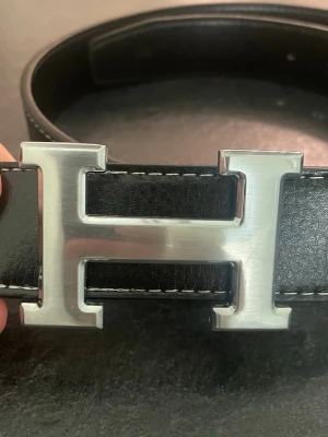 Hermmes belt - Säljer ett svart bälte i skinn med en stor, silverfärgad H-formad metallspänne. Bältet har vit kontrastsöm längs kanterna och flera hål för justering. Perfekt accessoar för att lyfta vilken outfit som helst.