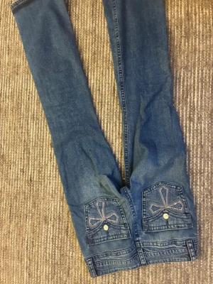 Blå bootcut jeans från Gina Tricot - Snygga blå jeans från Gina Tricot med bootcut-snitt och klassisk femficksdesign. Baksidan har coola fickor med broderade detaljer och knappstängning. Jeansen har dragkedja och knapp framtill, och är gjorda i mjukt bomullsmaterial.