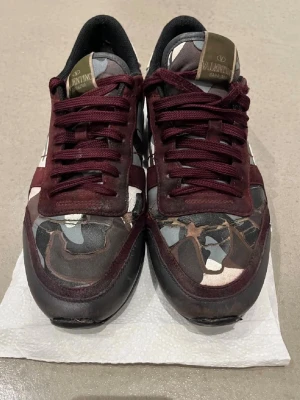 Valentino Rockrunner sneakers vinröd - Säljer ett par Valentino Rockrunner sneakers med vinröda detaljer och coolt camouflage-mönster i grått, svart och vitt. Skorna har snörning, rund tå och platt sula. Märkeslogga på plösen och paneler i skinn och textil. Perfekt för dig som gillar statement-skor.