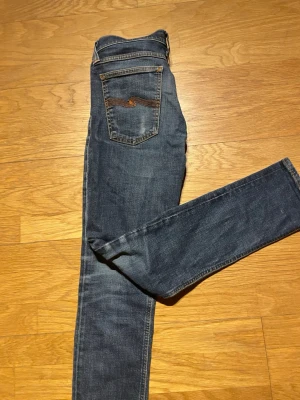 Blå jeans från Nudie Jeans, W31 L32 - Säljer ett par snygga blå jeans från Nudie Jeans i modellen Skinny Lin. De har klassisk femficksdesign, orangea sömmar och Nudie-logga på bakfickan. Jeansen är i stretchigt denim och har skinny passform med låg midja. 