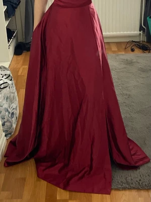 Vinröd balklänning med öppen rygg - Säljer en vinröd långklänning med snygg snörning i ryggen och öppen rygg. Den är aldrig använd och har prislapp. Klänningen har en elegant, vid kjol och är tillverkad i ett glansigt material som ger en lyxig känsla. Två fickor med en slit vid benet. Perfekt för speciella tillfällen och bal💕