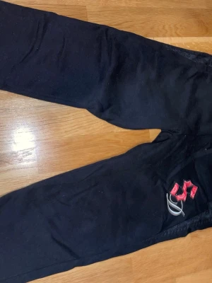 Svarta mjukisbyxor från supply&demand - Svarta mjukisbyxor med en unik paisley-detalj längs benslutet och ett broderat rött mönster på ena benet. Byxorna har en avslappnad passform och är gjorda i mjukt bomullsmaterial, perfekta för chill dagar. Snyggt broderi ger extra stilpoäng.