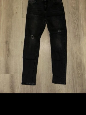 Svarta skinny jeans med slitningar - Svarta jeans med skinny passform och slitningar på båda knäna. Klassisk femficksmodell med smal siluett och normal midja. Jeansen är tillverkade i denim och har en mörk tvätt som ger en cool look.