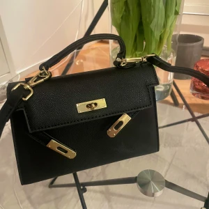 Svart handväska med gulddetaljer - Snygg svart handväska med stilrena gulddetaljer och struktur i materialet. Väskan har både handtag och axelrem, vilket gör den enkel att bära på olika sätt. Klassisk och elegant design med rektangulär form och lock med vridspänne i guld.  Ps. Inget märke på väskan. Oanvänd