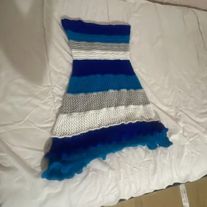 Blåvit stickad kortklänning med volang - Supersöt stickad kortklänning i blått, vitt och grått med flera olika stickade mönster och volangkant nertill. Klänningen är ärmlös och har en figurnära passform. Perfekt för dig som är utomlands sommaren 