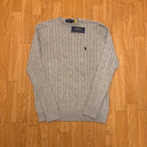 Grå stickad tröja Polo Ralph Lauren - Snygg grå stickad tröja från Polo Ralph Lauren med klassisk rund hals och broderad logga på bröstet. Tröjan är långärmad och har ribbade muddar vid ärmslut och nederkant. Perfekt för dig som gillar stilrena och tidlösa plagg. Extreeemt bra kvalite på dessa passa på och köp innan de tar slut!!!