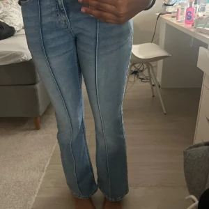 Ljusblå bootcut jeans med detaljer - Säljer ett par ljusblå jeans med bootcut-modell och markerade sömmar längs benen. Snygga bakfickor med lock och dekorativa sömmar. Klassisk femficksdesign och normal passform. Perfekta för dig som gillar en retrovibe och vill sticka ut lite extra.