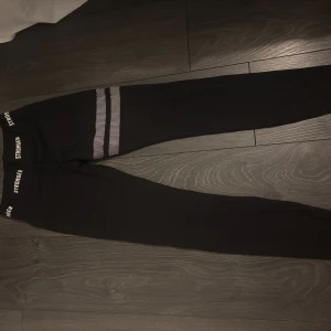 Svarta Stronger leggings med vita detaljer - Svarta leggings från Stronger med vita logotyper i midjan och två vita randiga detaljer på ena benet. Tight passform och stretchigt material som passar perfekt till träning. Snygg och sportig look med hög midja. Som man ser på sista bilden har det försvunnit lite på ränderna💕