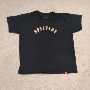 Svart Spitfire t-shirt i bomull - Svart t-shirt från Spitfire med tryckt logga i gult och rött på bröstet. Klassisk passform med korta ärmar och rund hals. Tillverkad i 100% bomull, perfekt för en avslappnad streetwear-look.